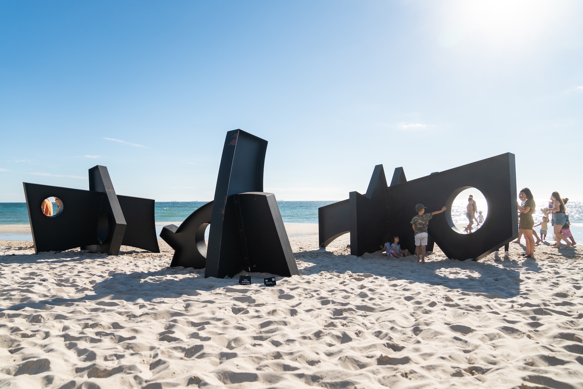 Large black geometric sculptures on a sandy beach with people interacting and the ocean in the background.