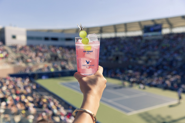 Person holding a pink drink garnished with grapes at a tennis match in a large stadium.