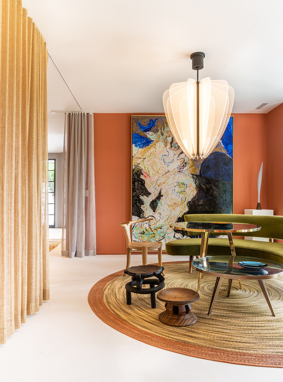 Modern living room with round rug, green sofa, abstract art, large pendant light, and wooden stools.