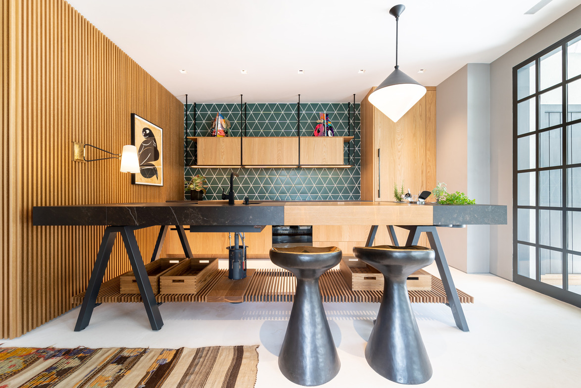 Modern kitchen with geometric tile backsplash, wooden cabinets, abstract art, black counters, and unique stools.