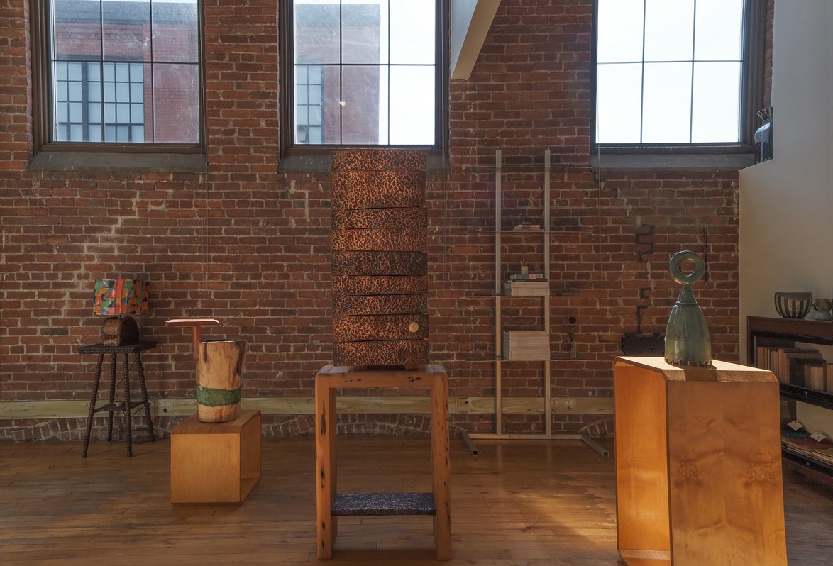 Art gallery space with brick walls featuring wooden sculptures and abstract art pieces on display in warm natural lighting.