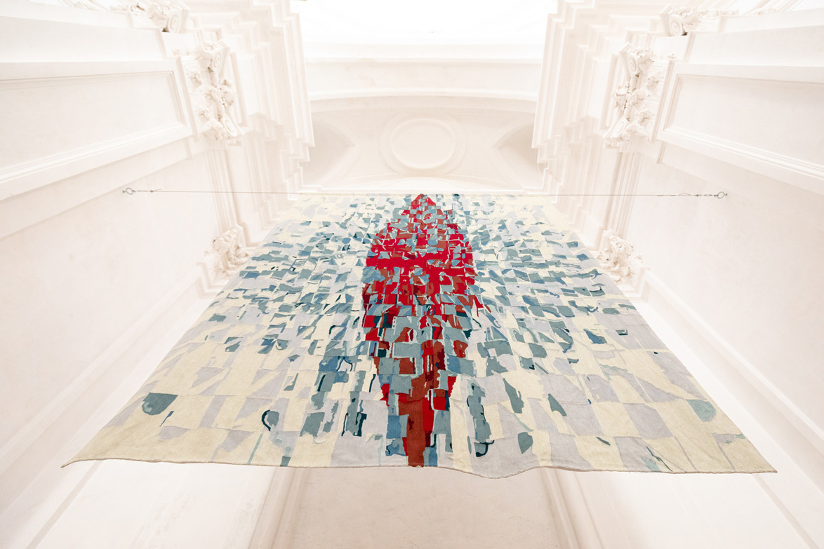 Large abstract fabric art installation with red and blue geometric patterns suspended in ornate white architectural space.