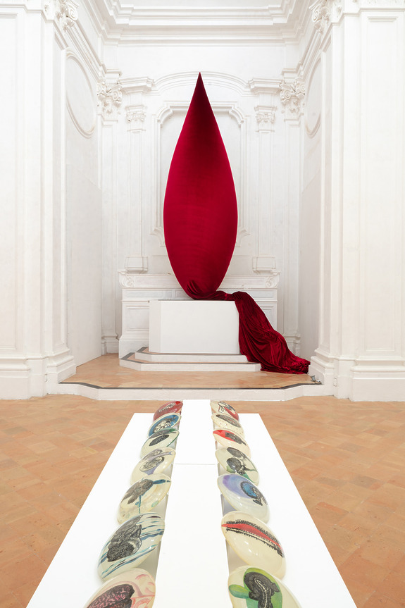 Large red sculptural drop with vibrant fabric in a white ornate room, accompanied by a series of abstract objects on the floor.