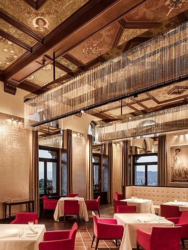 Elegant restaurant interior with ornate ceiling, luxurious chandeliers, red chairs, and large windows offering scenic views
