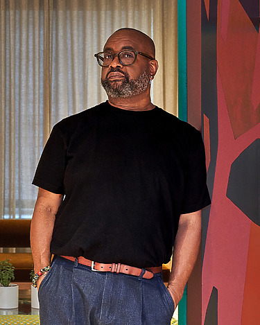 Man wearing a black t-shirt and glasses standing indoors near colorful wall art and window with sheer curtains.