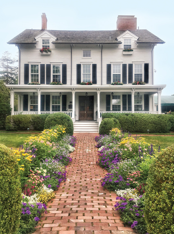 Victorian-style house with a brick pathway and lush flower garden in the foreground.