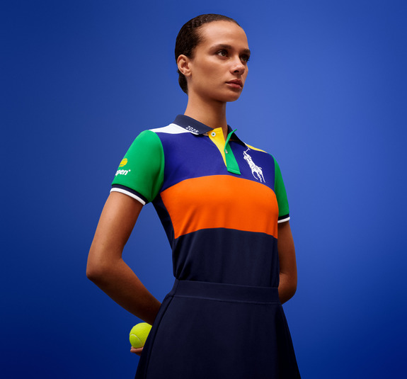 Person in multicolored polo shirt holding a tennis ball against a blue background.