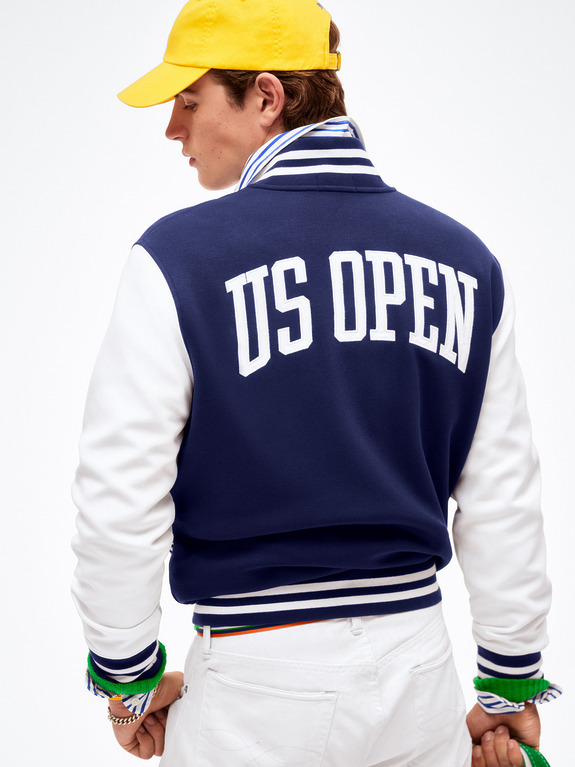 Person wearing a US Open varsity jacket, striped collar shirt, yellow cap, and white pants, viewed from the back.
