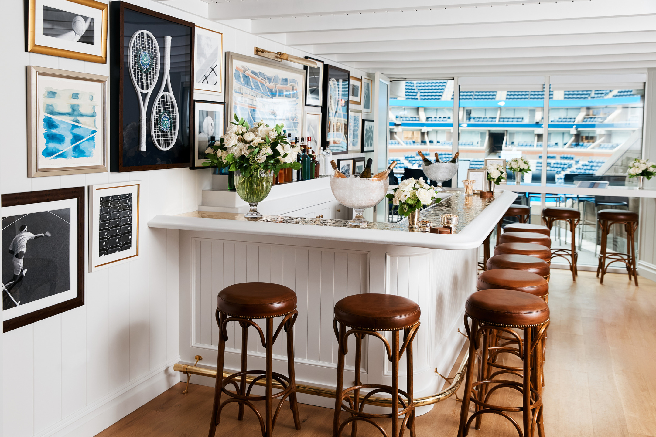 Stylish bar area with brown stools, decorative art, white counter, and floral arrangements overlooking a sports stadium.