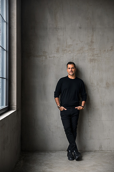 Man in black outfit standing against a concrete wall next to a large window, hands in pockets, and casually leaning.