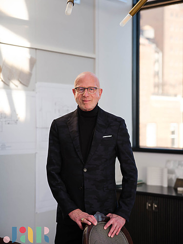 Man in a dark suit standing in a modern office with architectural drawings on the wall behind him