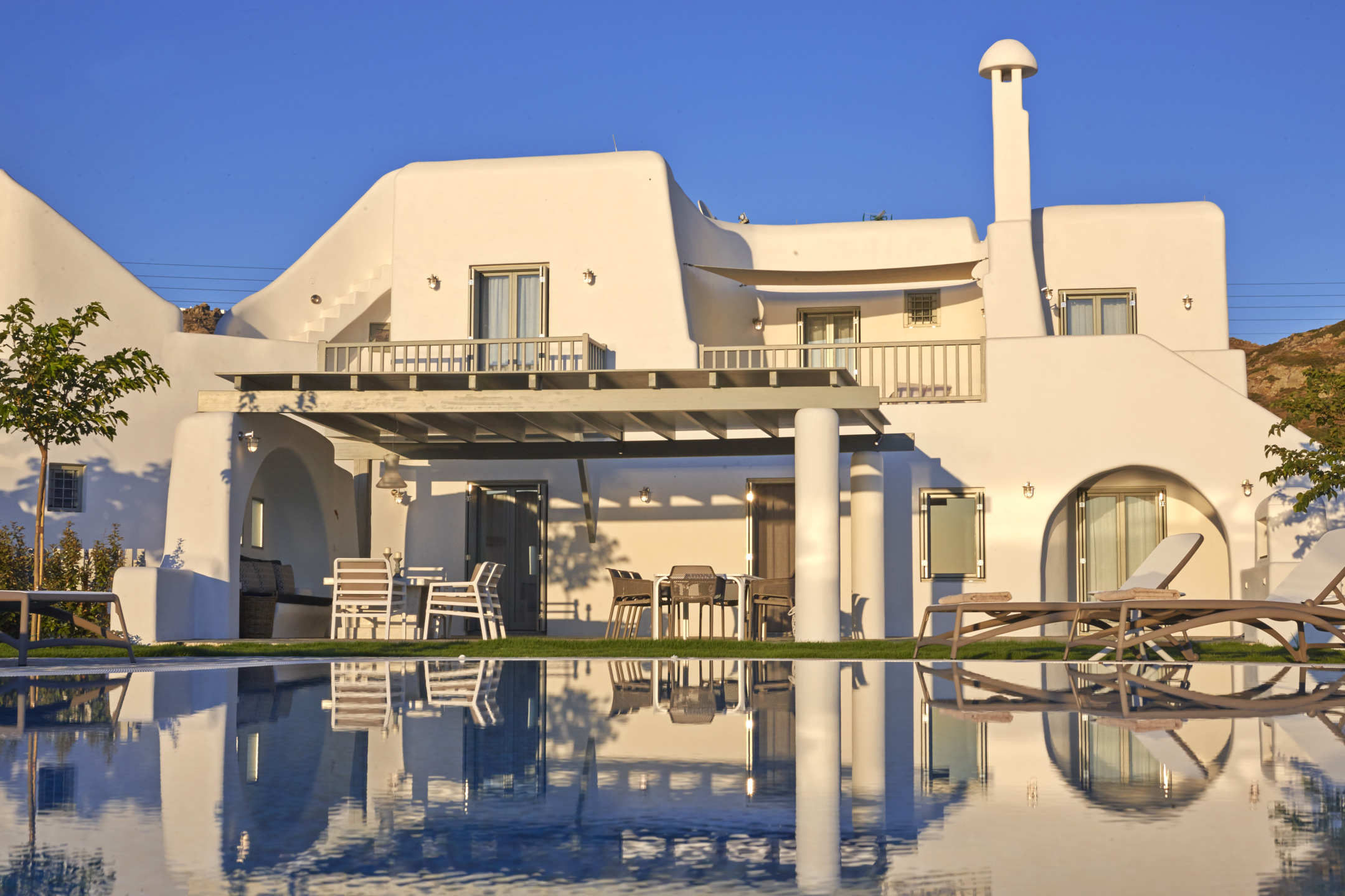 Modern white villa with a pool, surrounded by clear blue sky and lush greenery, reflecting in the water.