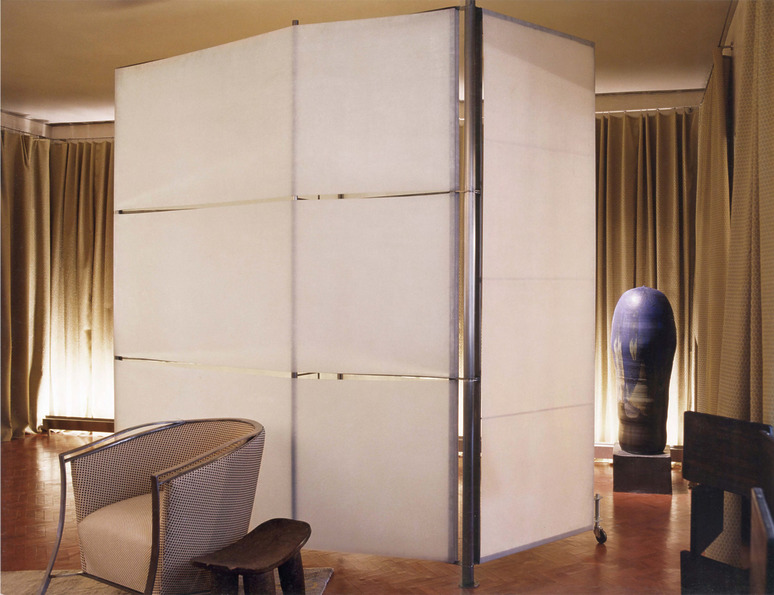 Room divider with white panels in a modern living room with wooden chair, small stool, and blue decorative vase.