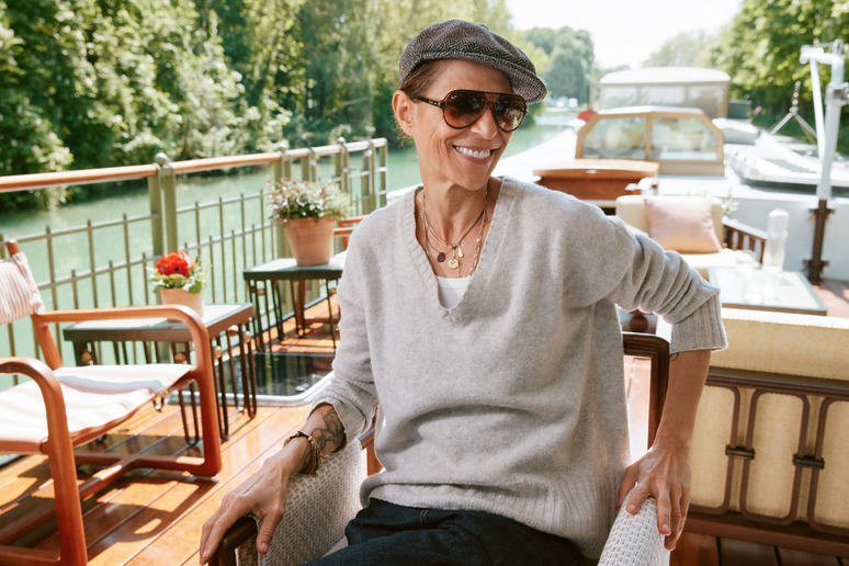 Smiling person wearing sunglasses and hat sitting on a boat deck with chairs and greenery in the background.