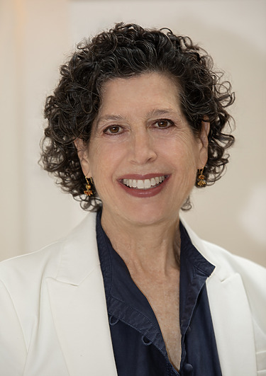 Smiling person with short curly hair wearing a white jacket and navy shirt.