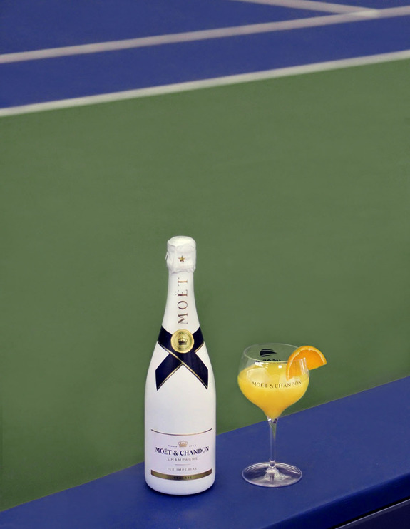 Champagne bottle and orange cocktail on tennis court sideline.