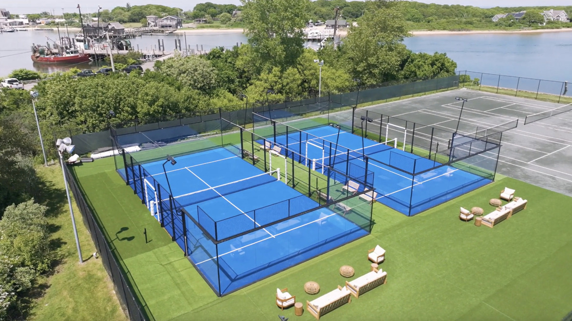 Aerial view of paddle tennis courts with bright blue surfaces near a waterfront and greenery.