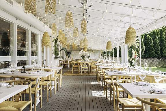 Outdoor restaurant patio with wooden tables, chairs, and hanging wicker lights, surrounded by greenery and set for dining.