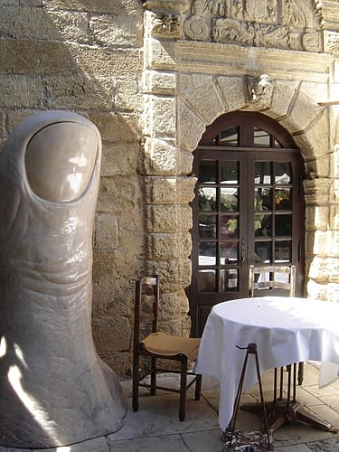 Stone building with large thumb sculpture, a wooden chair, and a round table covered with a white cloth in the foreground.