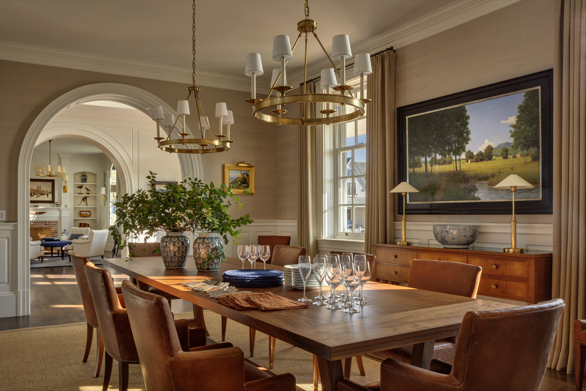 Elegant dining room with wooden table, leather chairs, chandeliers, artwork on wall, and arched doorway leading to another room.