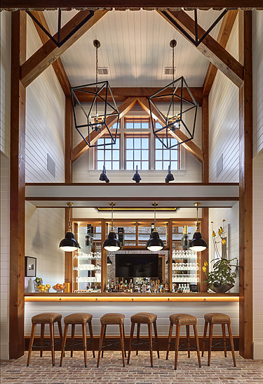 Rustic home bar with wooden stools, geometric chandeliers, and shelves lined with bottles under a high vaulted ceiling.