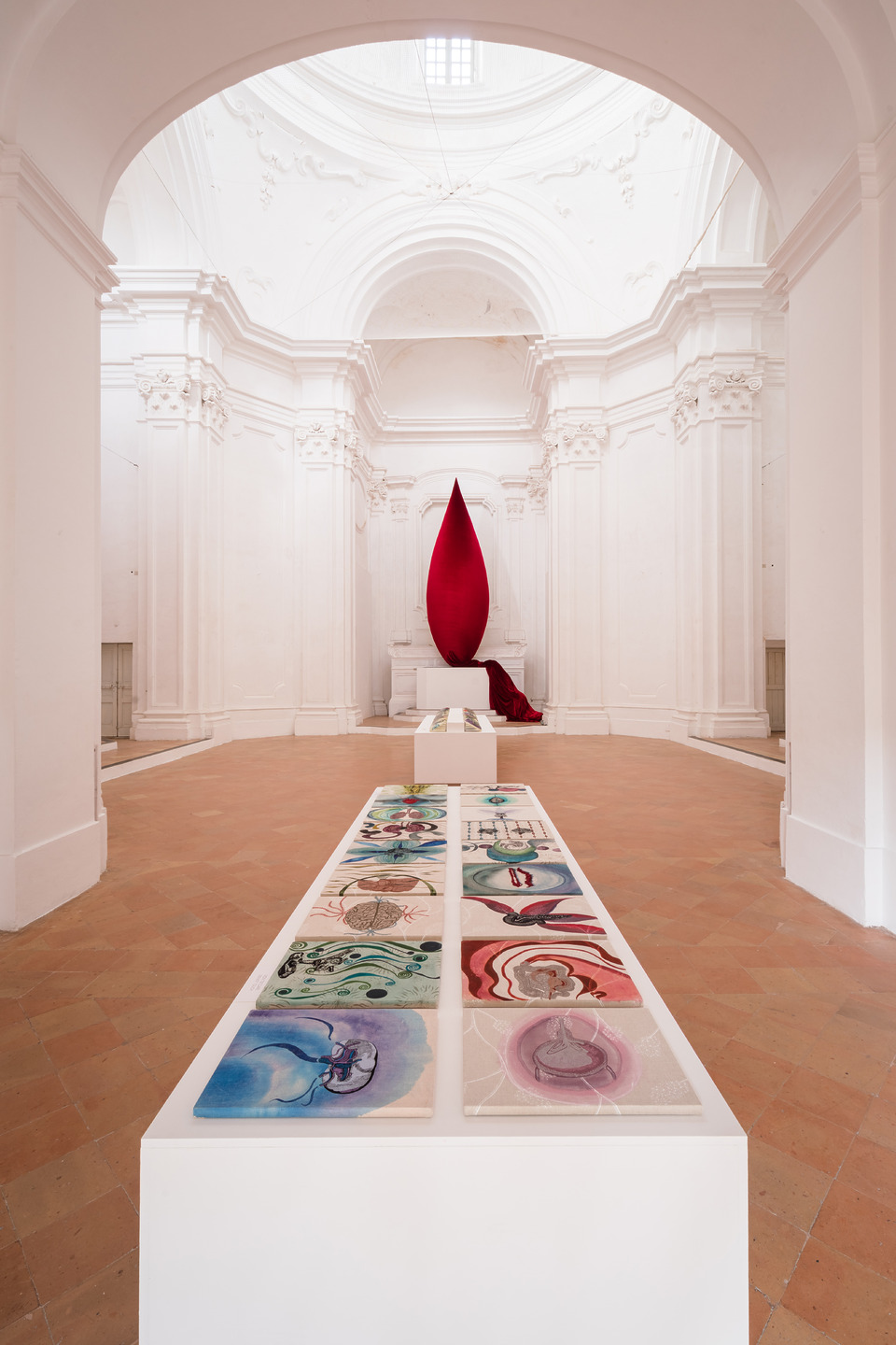 Contemporary art installation in a bright gallery with a red sculpture and colorful paintings on a central display.