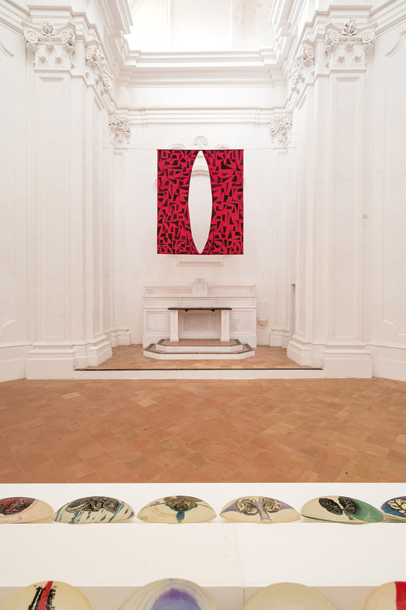 Art installation in a white-walled gallery, featuring a red and black abstract painting on the wall and circular plates on the floor.