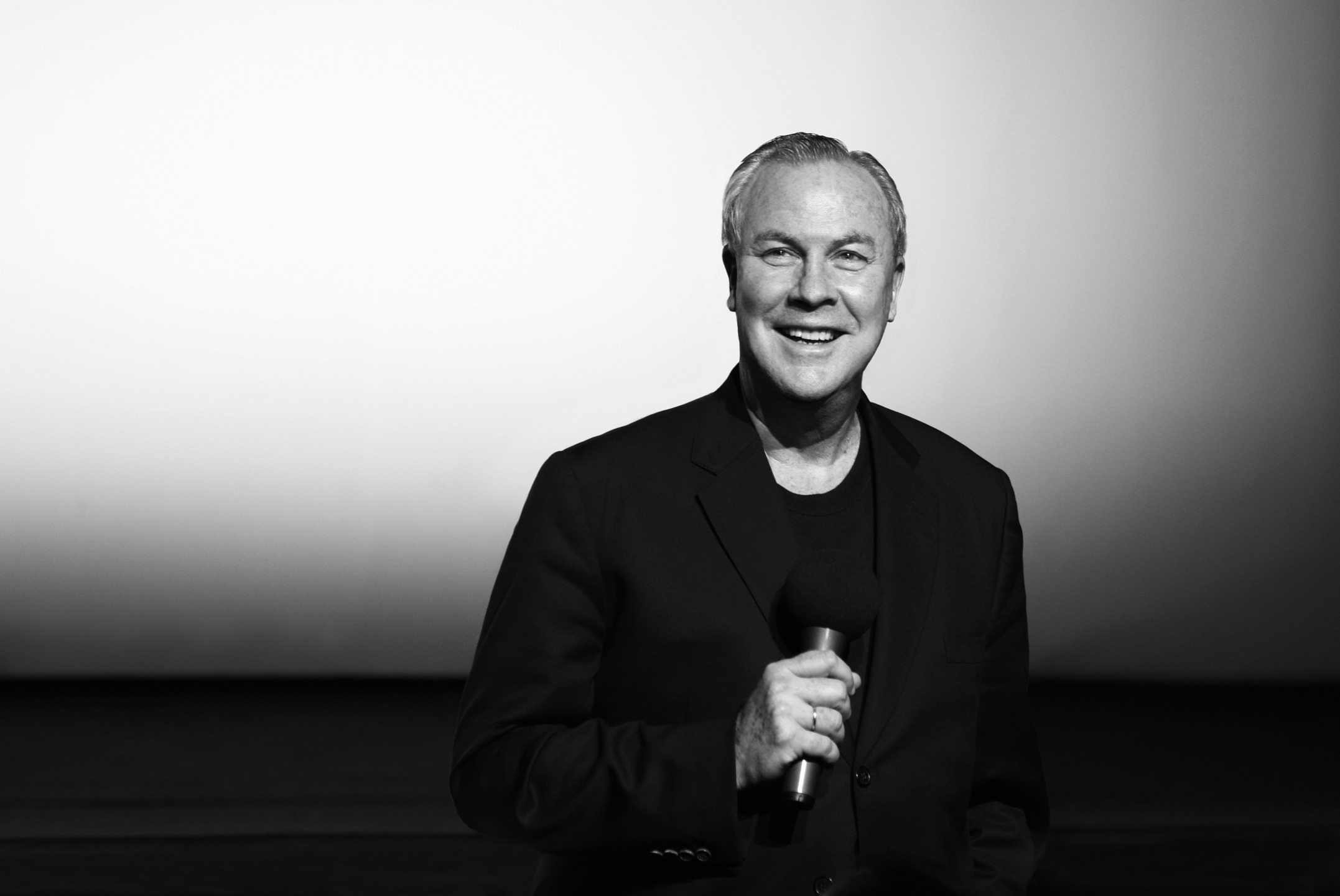 Man in a black suit holding a microphone, standing on a stage with a neutral background.