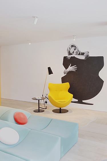 Modern living room with yellow chair, colorful sofa, and black-and-white wall art of a woman peeking from behind a shape.