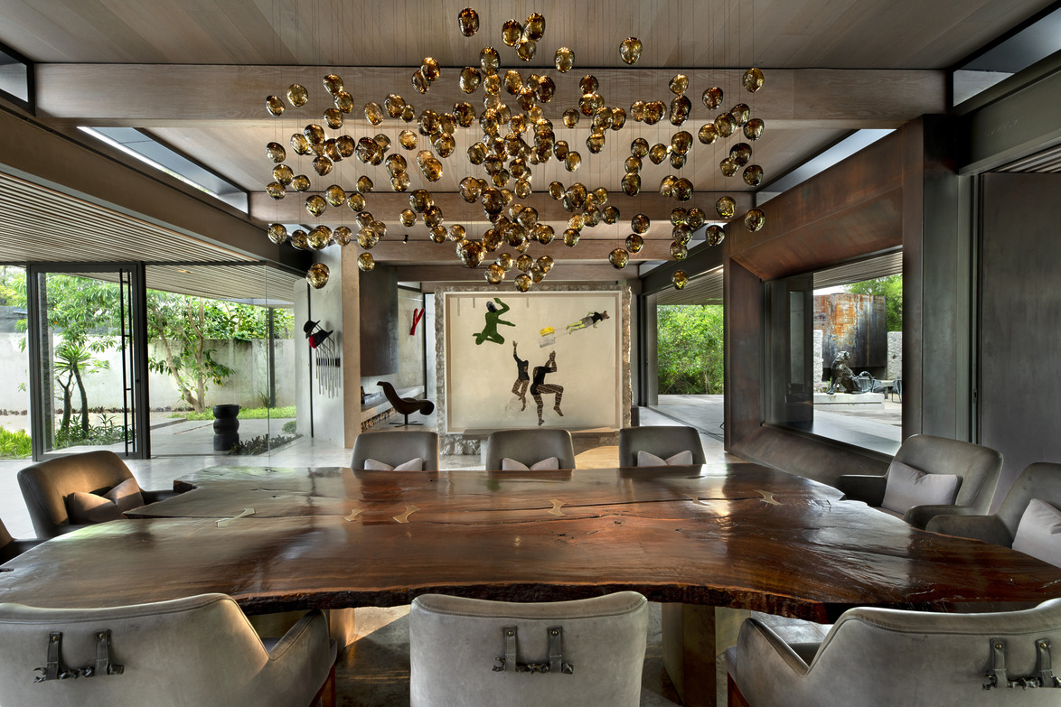 Modern dining room with large wooden table, unique chandelier, and contemporary art on walls, surrounded by windows.
