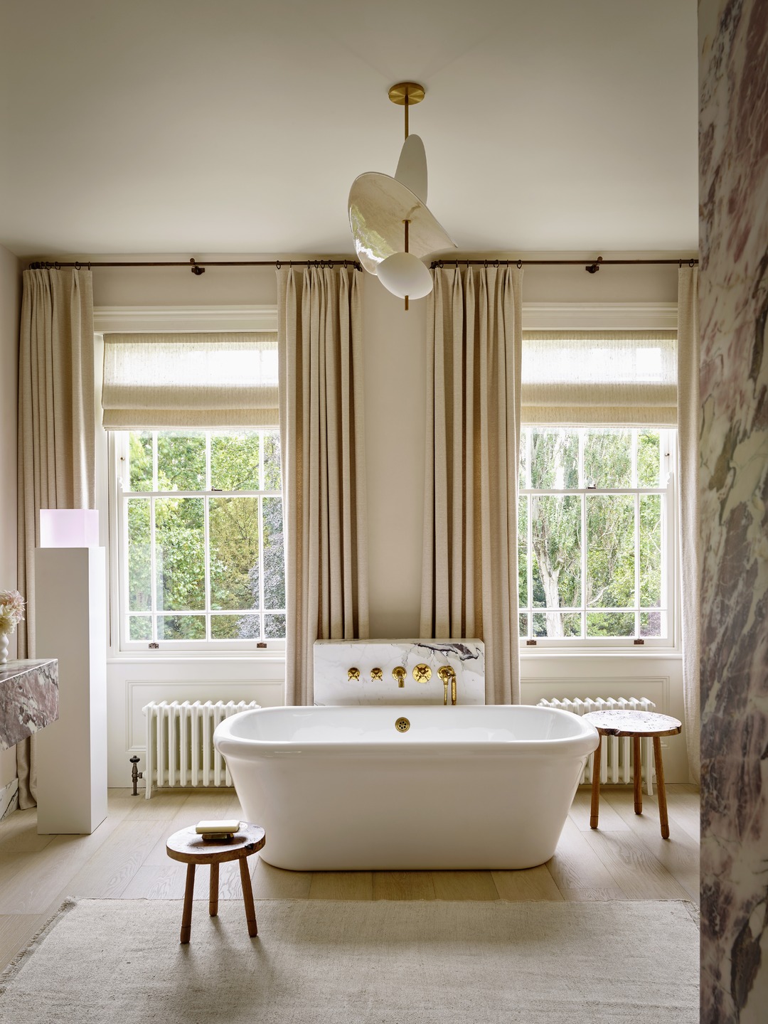 Elegant bathroom with a freestanding tub, tall windows with drapes, wooden stools, and a modern ceiling light fixture.