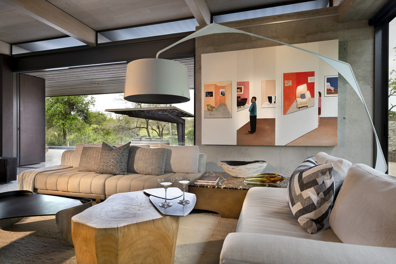 Modern living room with large abstract painting, sleek furniture, and unique lighting, overlooking lush outdoor scenery.