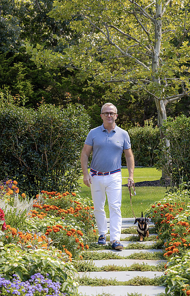 Man walking a small dog along a garden path surrounded by vibrant flowers and greenery