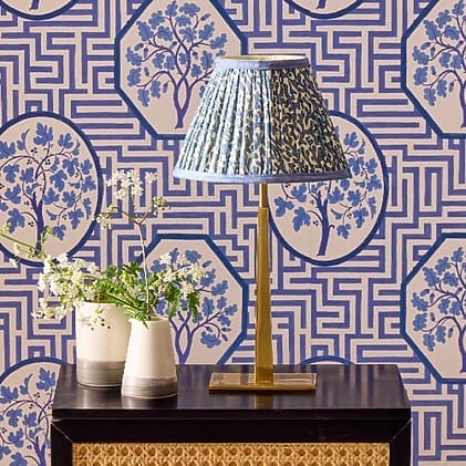 Table lamp with blue shade on dark wood table, set against a geometric blue and white floral wallpaper background.