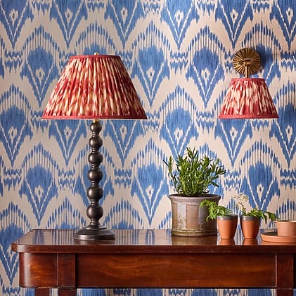 Table with red lampshade, blue patterned wallpaper, and plants in clay pots.