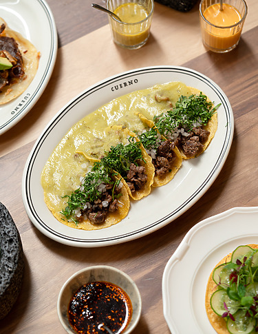 Four tacos with various toppings on a white plate, surrounded by dipping sauces and drinks on a wooden table.