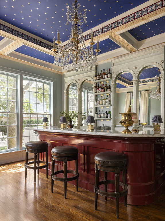 Luxurious home bar with red counter, elegant chandelier, and star-patterned ceiling, surrounded by large windows.