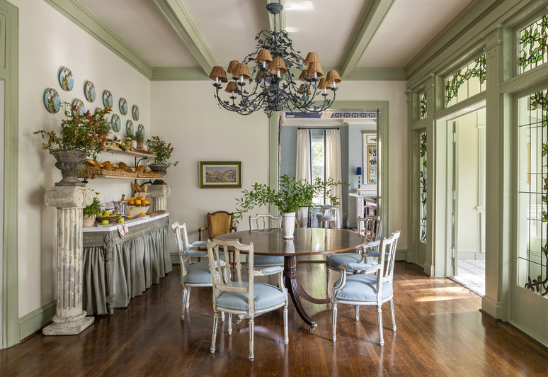 Elegant dining room with a round table, blue chairs, chandelier, and decor items on a shelf against a wooden floor backdrop.