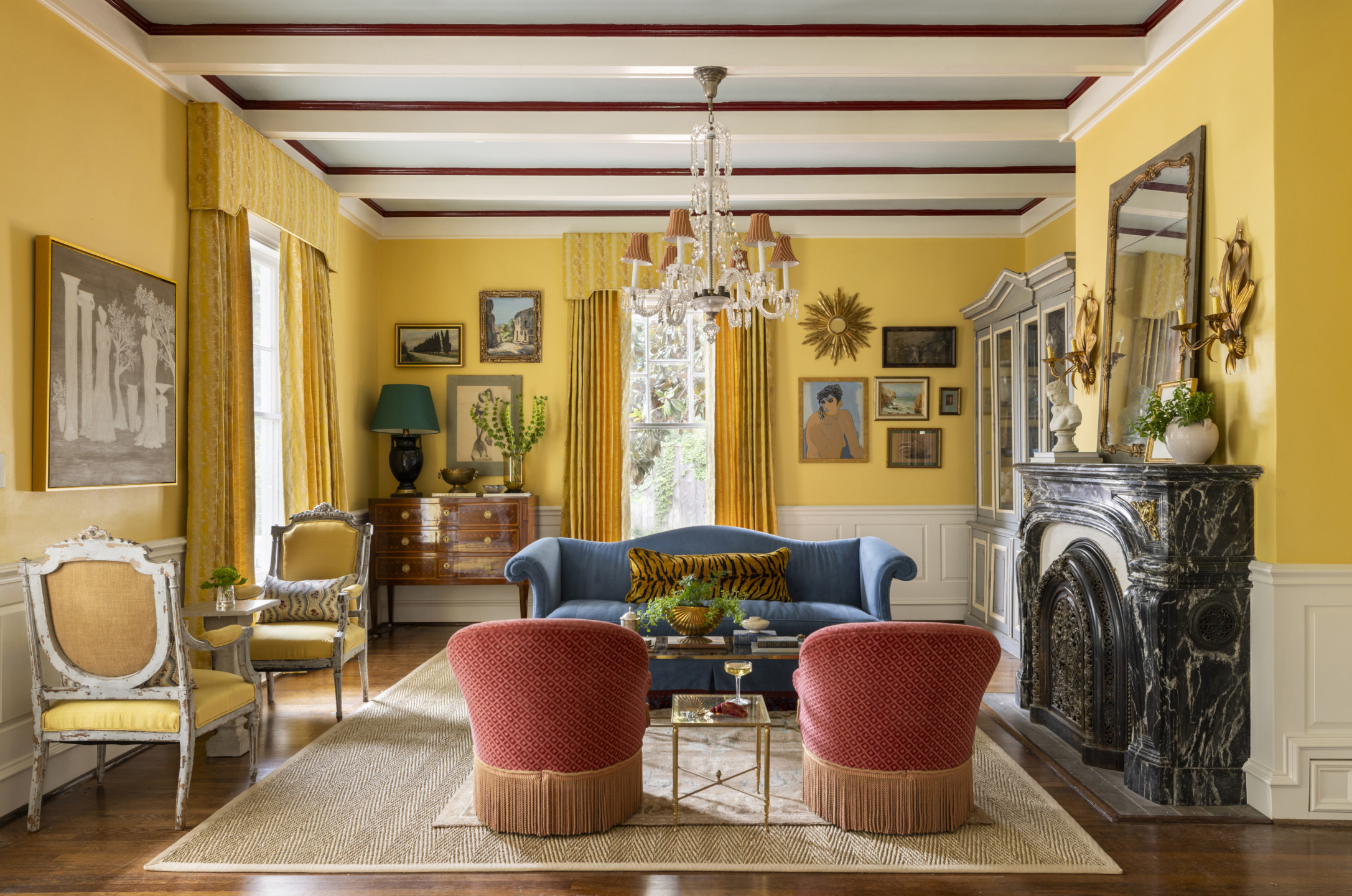 Elegant living room with yellow walls, vintage furniture, blue sofa, red chairs, chandelier, and a decorative fireplace.