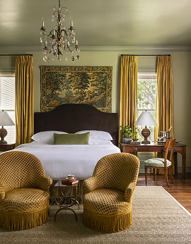 Elegant bedroom with chandelier, tapestry, and mustard yellow armchairs, featuring a dark brown headboard and classic decor.