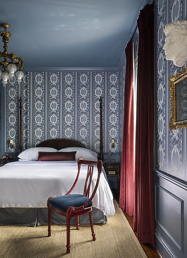 Elegant vintage bedroom with ornate wallpaper, blue ceiling, red drapes, antique bed, and a decorative wooden chair.