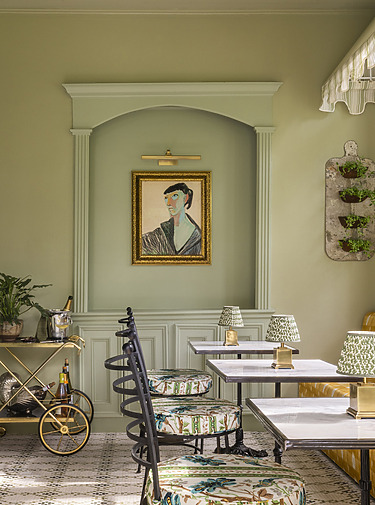 Elegant cafe interior with patterned chairs, small tables, a gold painting, and a vintage bar cart against a light green wall.