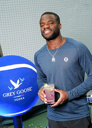 Man smiling and holding a cocktail next to a large Grey Goose vodka tennis ball decoration indoors.