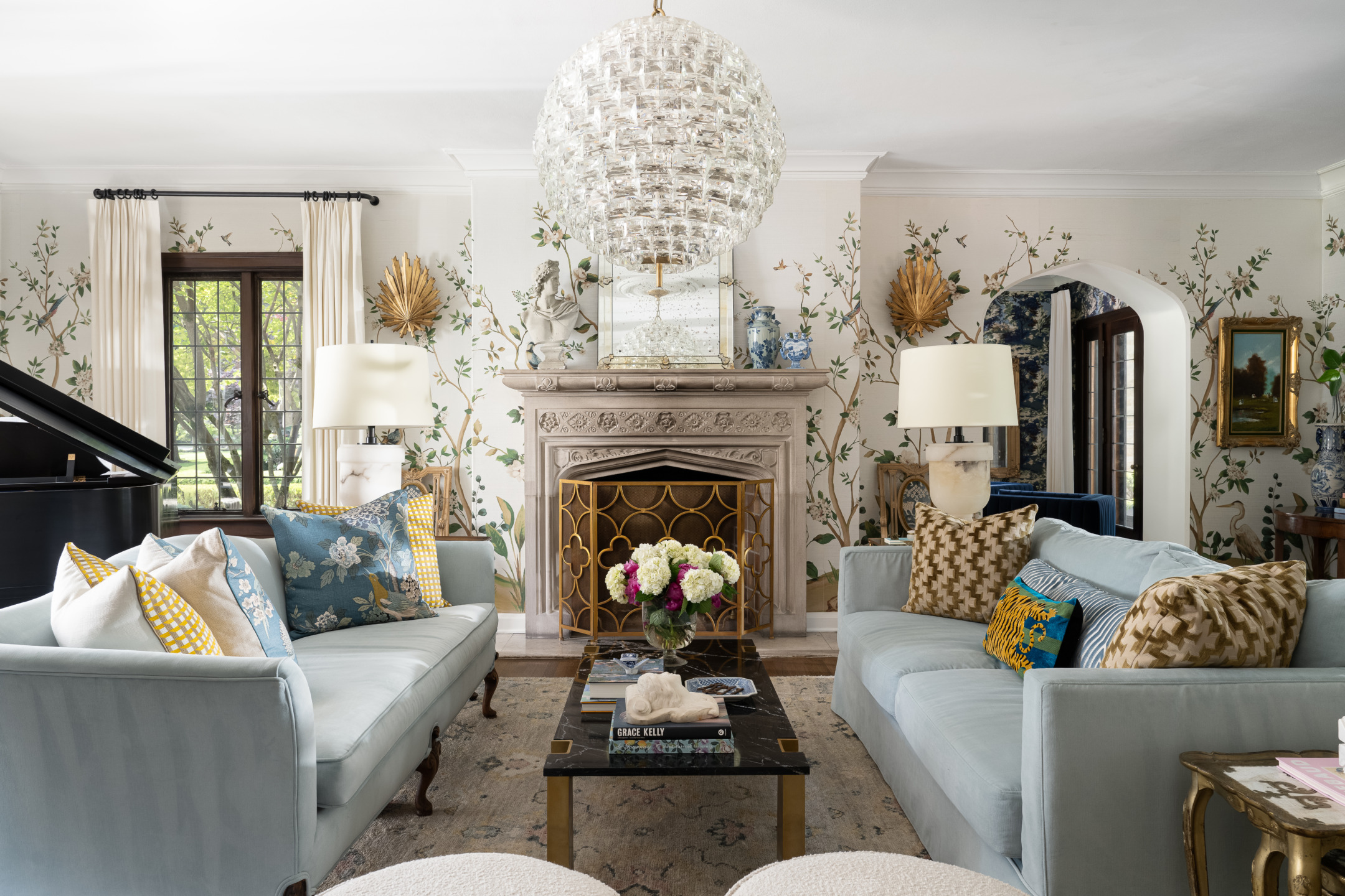 Elegant living room with floral wallpaper, light blue sofas, decorative fireplace, chandelier, and a grand piano in the corner.