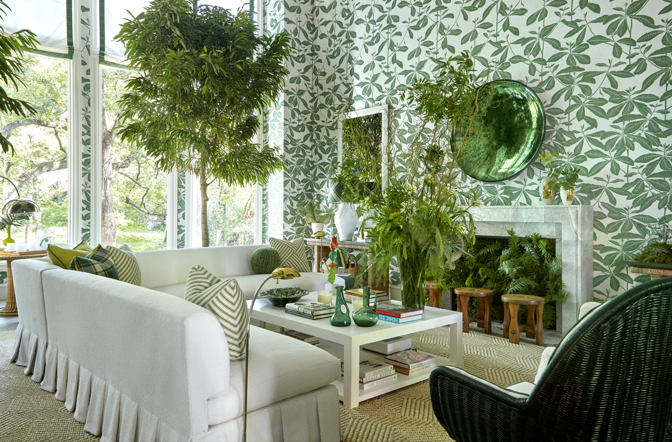 Bright living room with leafy wallpaper, white sofa, green plants, wicker chair, and a round mirror above the fireplace.