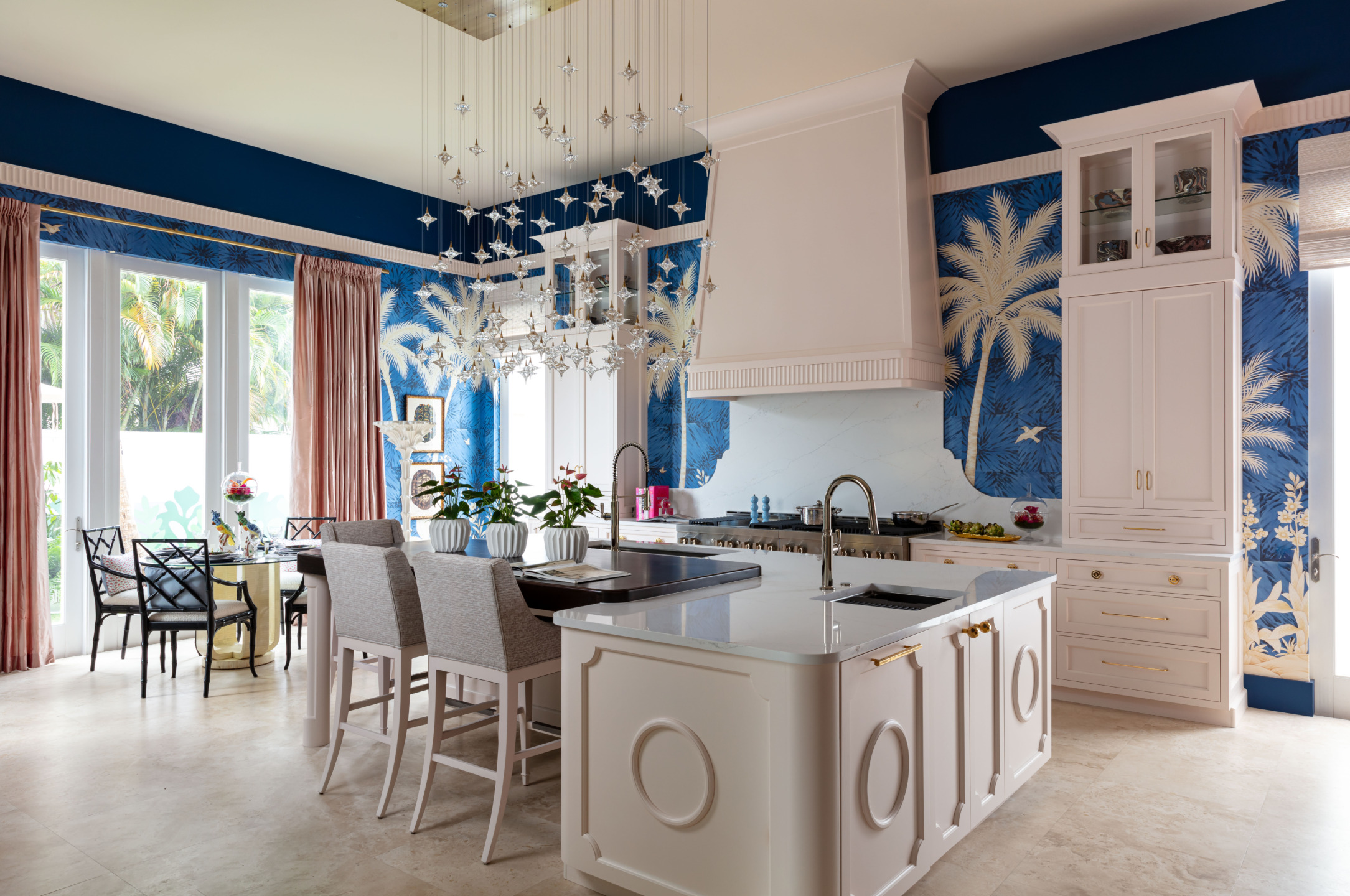 Luxurious kitchen with white island, blue tropical wallpaper, elegant lighting, and large windows overlooking a garden.