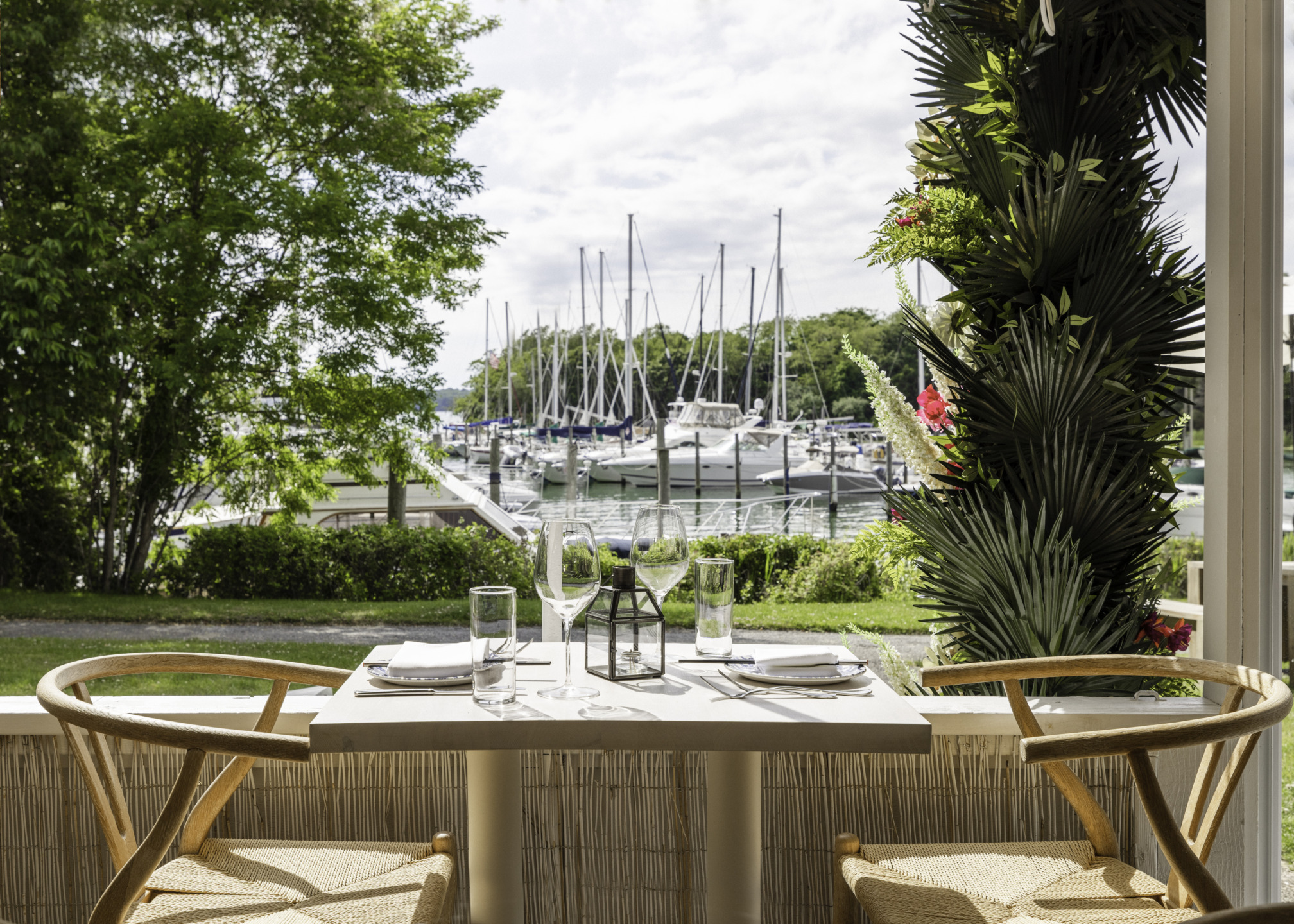 Outdoor dining table set for two overlooking a marina with sailboats and lush greenery in the background.