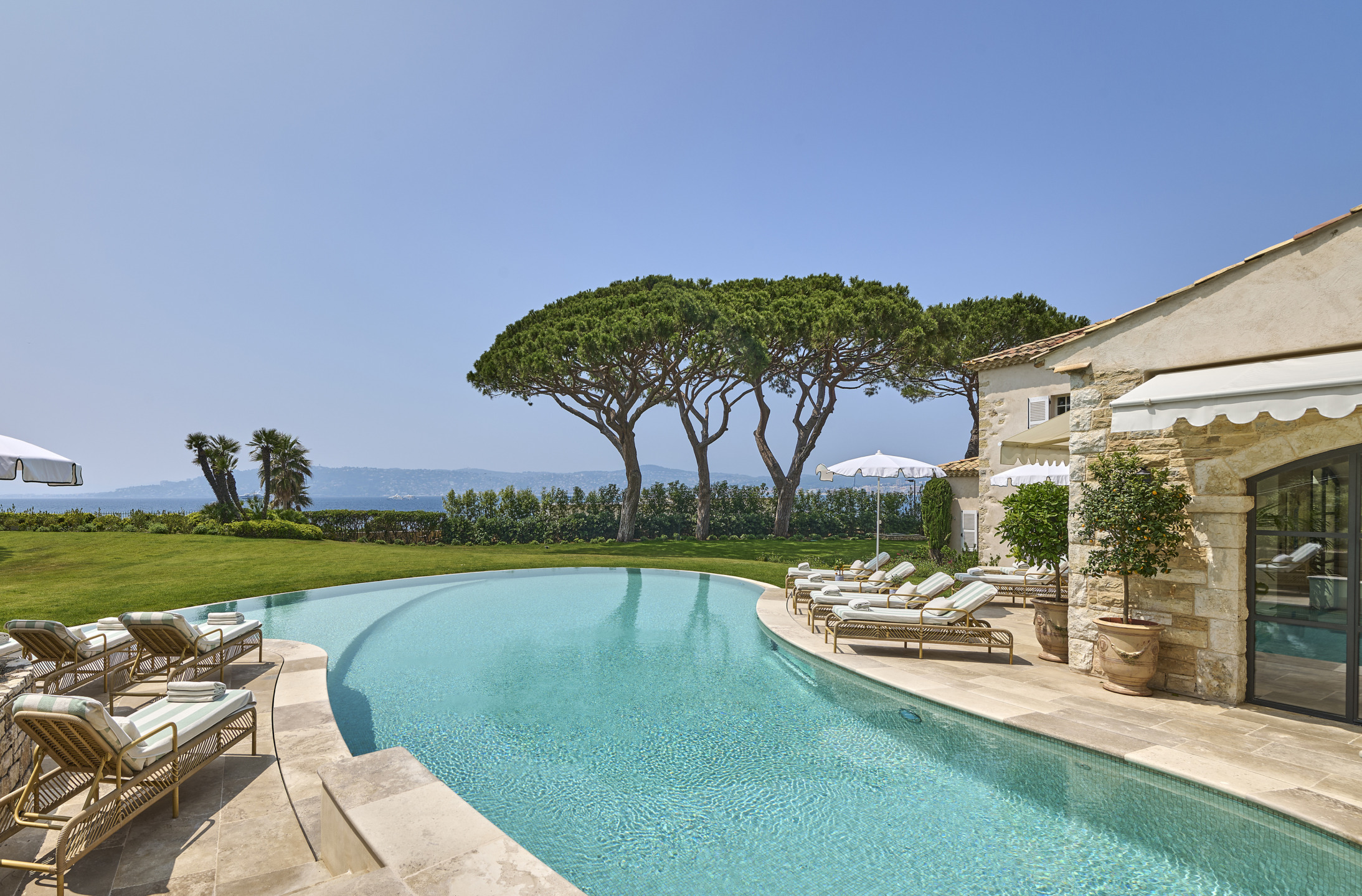 Luxury villa with curved pool, sun loungers, and umbrella. Lush greenery and trees in the background against a clear sky.