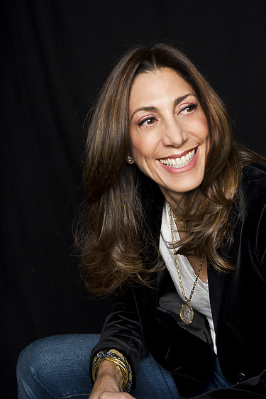 Smiling woman with long brown hair wearing a black jacket and a necklace against a dark background.