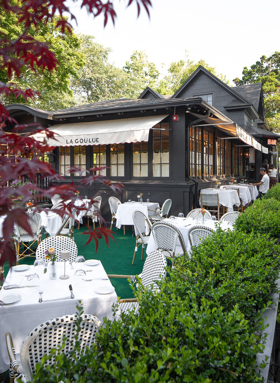 Outdoor dining area with white tables and chairs at a restaurant surrounded by greenery and a building in the background.